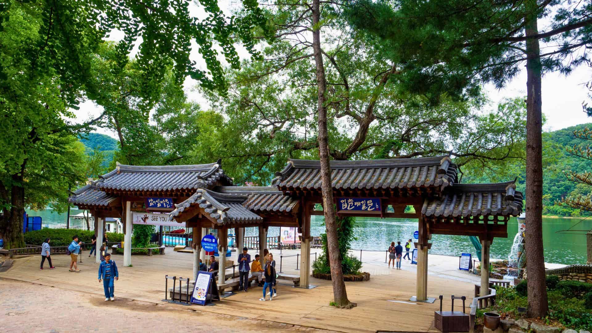 Traditional Korean wooden gates by a lakeside, surrounded by trees and people walking nearby.