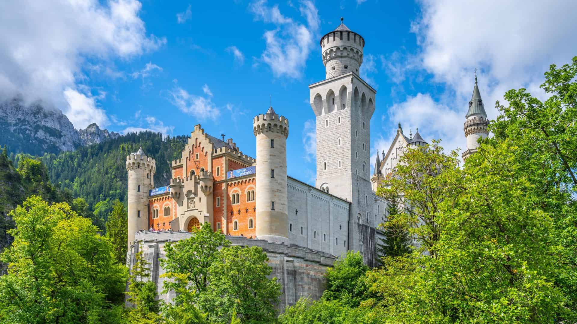 Neuschwanstein Castle rises above green trees with blue sky and mountains in the background.