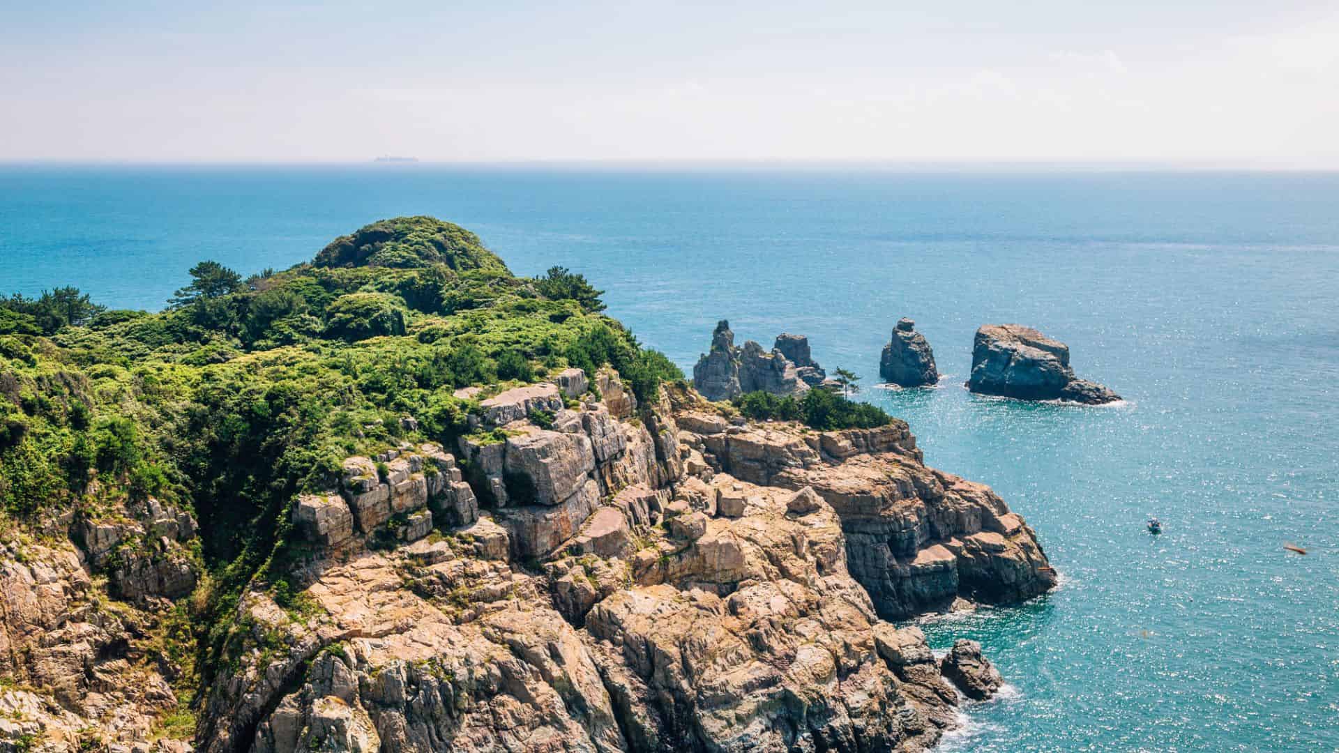 Rocky cliffs with green vegetation overlooking a calm blue sea, with rocks and islands in the water.