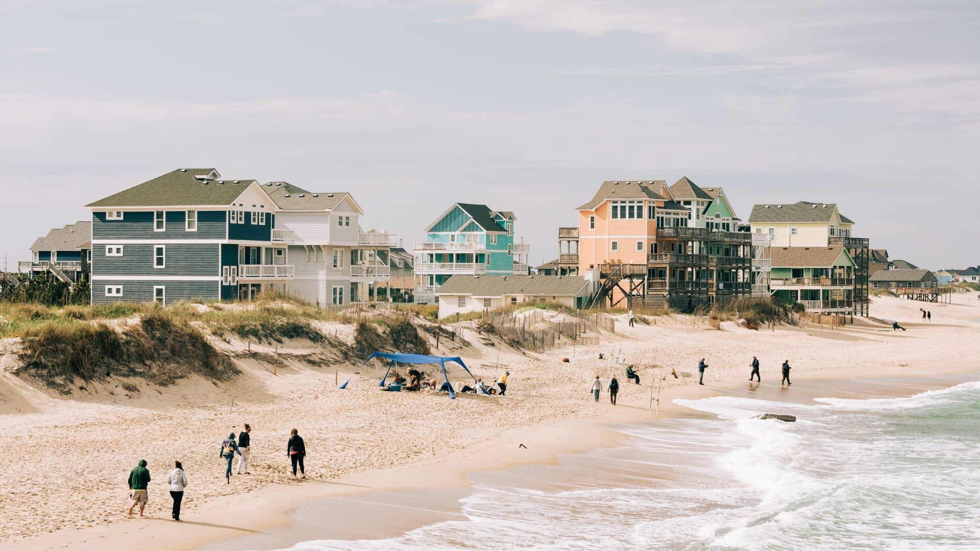 People walking along a sandy beach with colorful beach houses in the background under a partly cloudy sky.