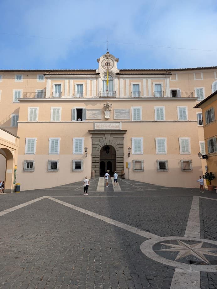 A large beige building with open windows, a clock, and people walking in a cobblestone courtyard—perfect for spending 4 days in Castel Gandolfo.