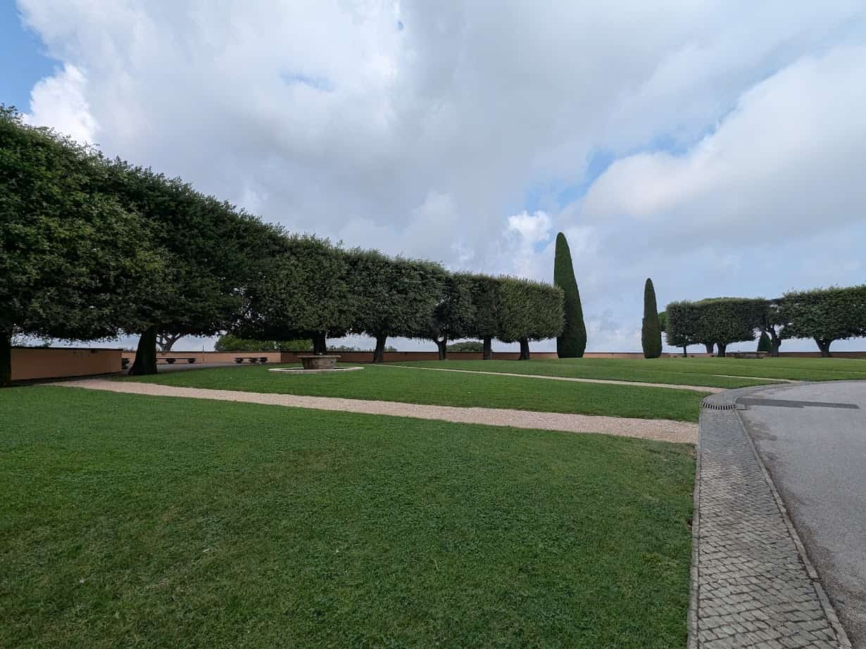A neatly trimmed lawn with rows of pruned trees and a paved walkway under a partly cloudy sky, perfect for enjoying 4 days in Castel Gandolfo.