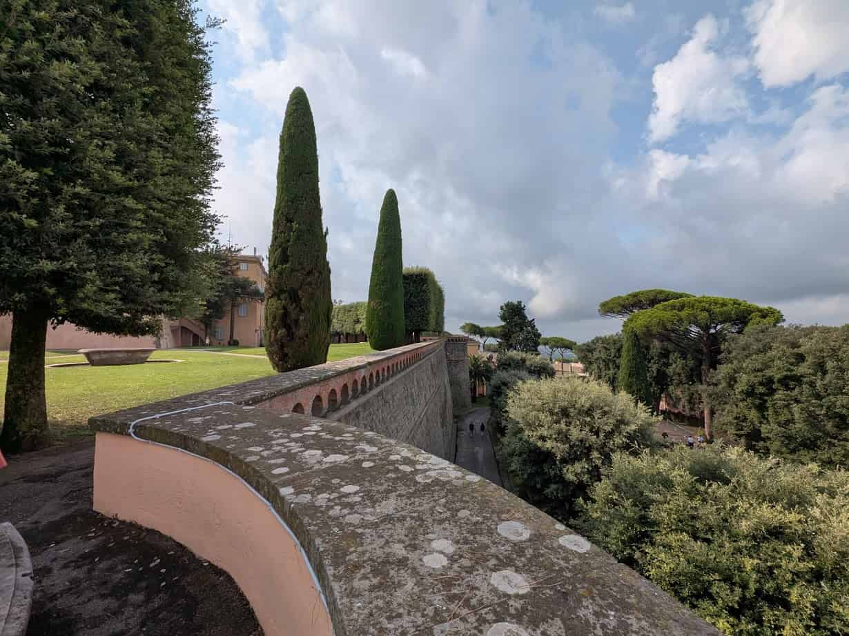 A stone wall and walkway lined with trees, with a grassy area and a cloudy sky in the background, capture the peaceful charm you might discover during 4 days in Castel Gandolfo.