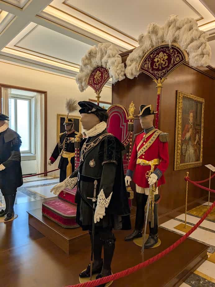 Historical royal costumes and uniforms displayed on mannequins beside ornate thrones and feathered canopies, evoke the grandeur one might experience during 4 days in Castel Gandolfo.