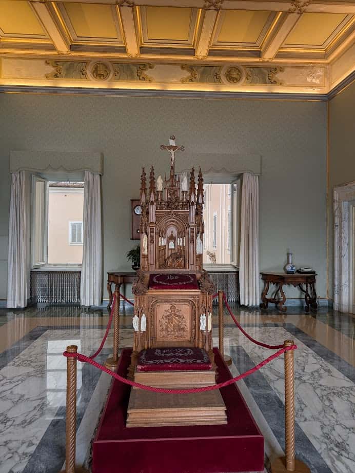 Ornate wooden throne with red velvet, roped off in a marble-floored, elegant room with high ceilings—just one of many regal sights to discover during 4 days in Castel Gandolfo.