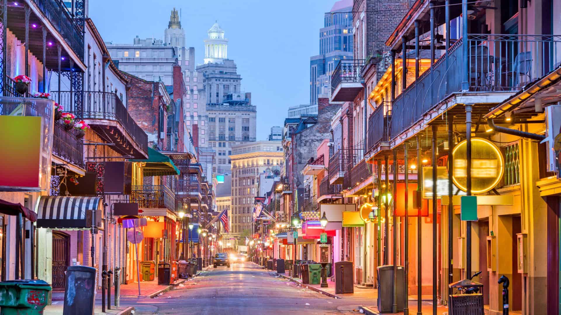 Colorful neon signs and historic buildings line a lively street in New Orleans at dusk.