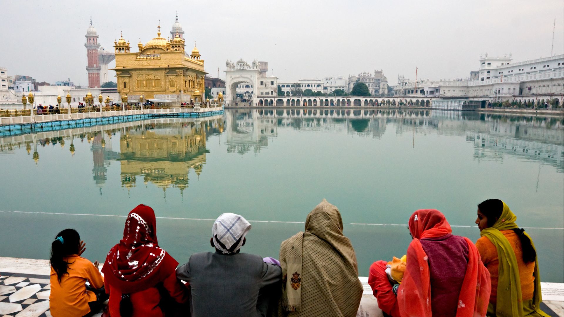 Six people in colorful clothing sit by the water, facing the Golden Temple in Amritsar, India.