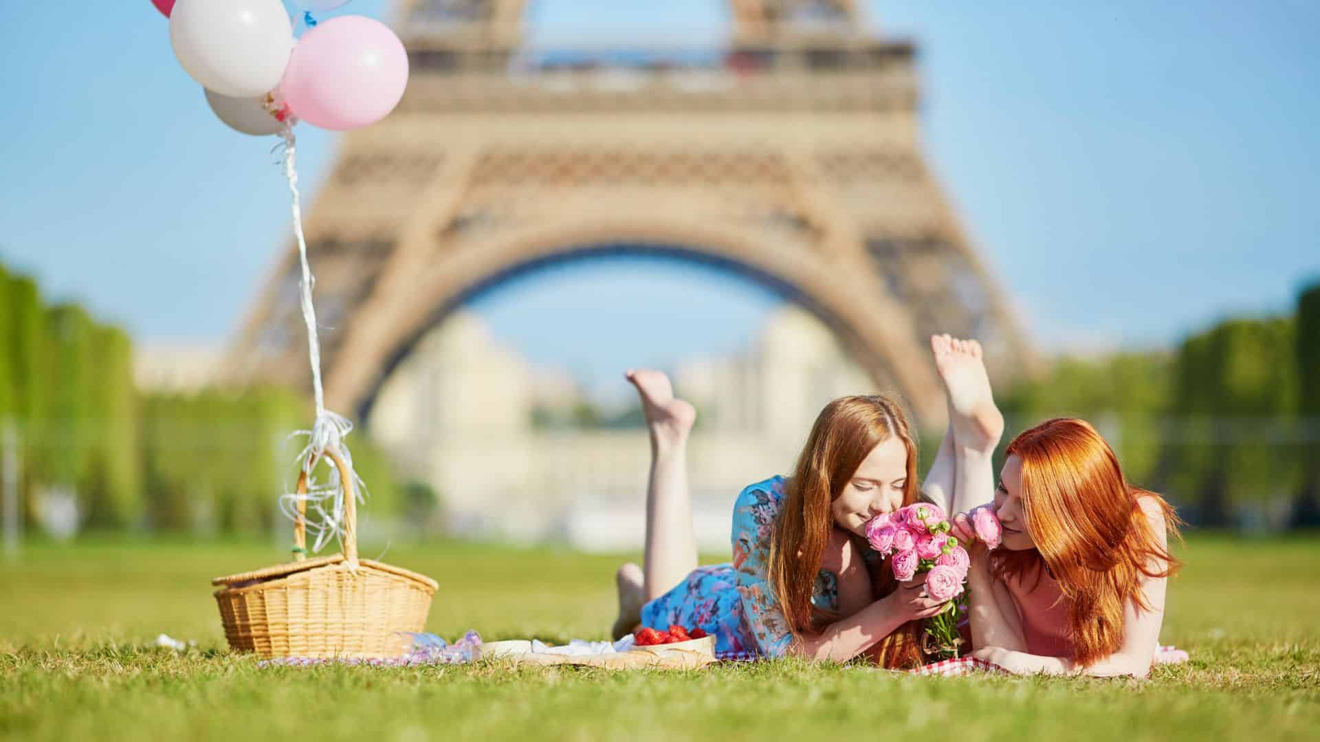 Two women lie on grass near a picnic basket, smelling flowers with the Eiffel Tower in the background.
