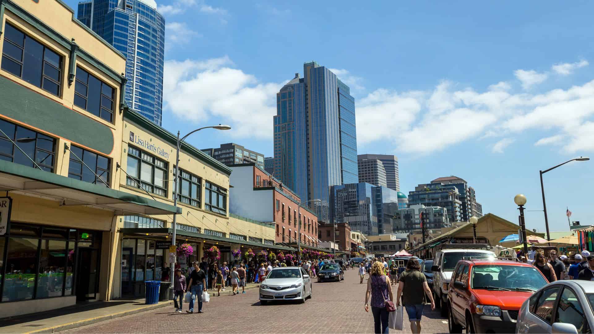 Busy city street lined with shops, people walking, tall buildings, and cars under a sunny blue sky.