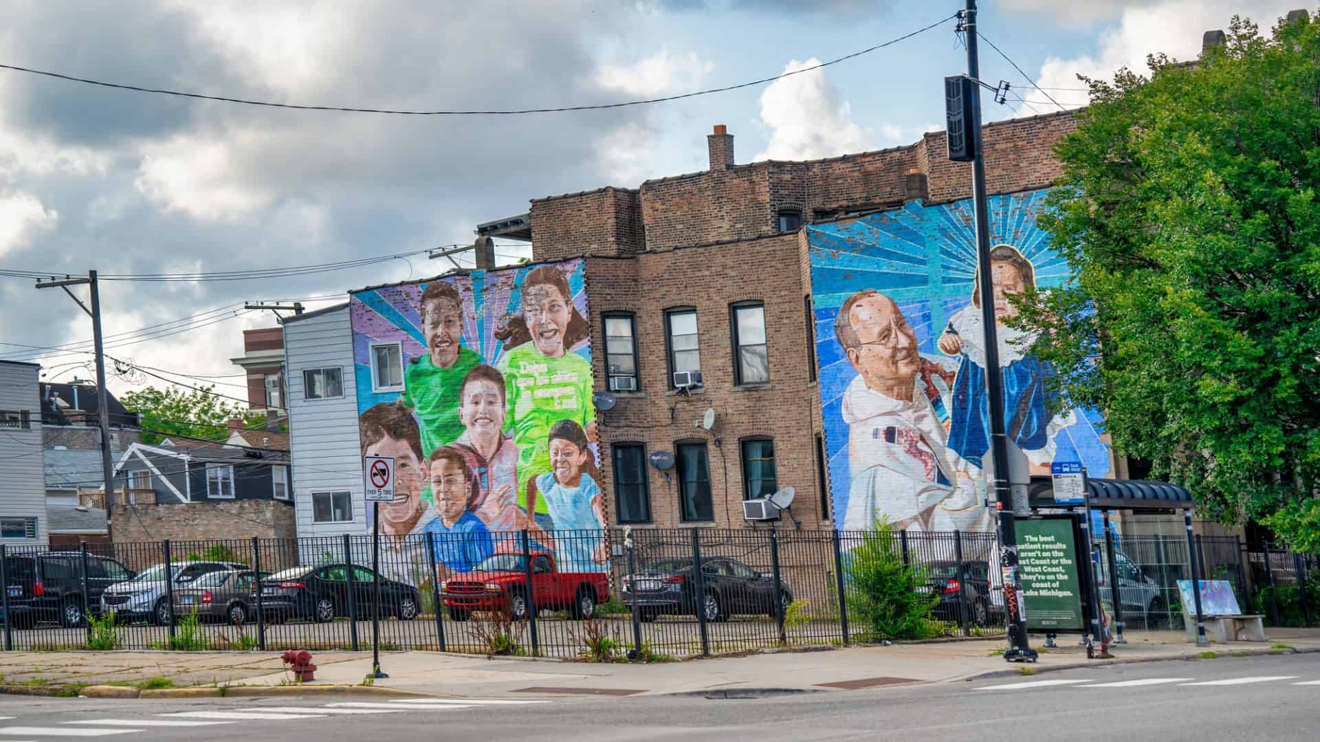 Colorful mural of smiling children and adults painted on the side of a brick building near a parking lot.