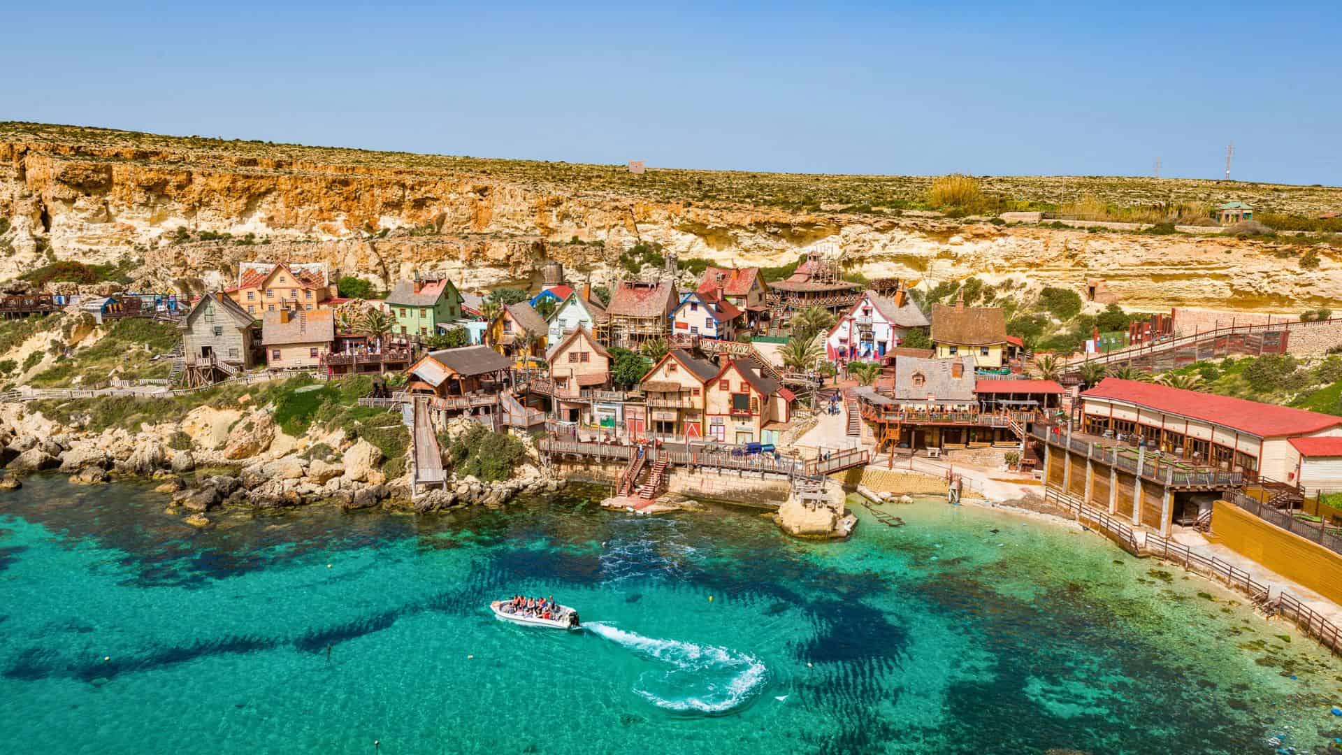 Colorful seaside village with quirky houses by clear turquoise water and a boat, surrounded by rocky cliffs.