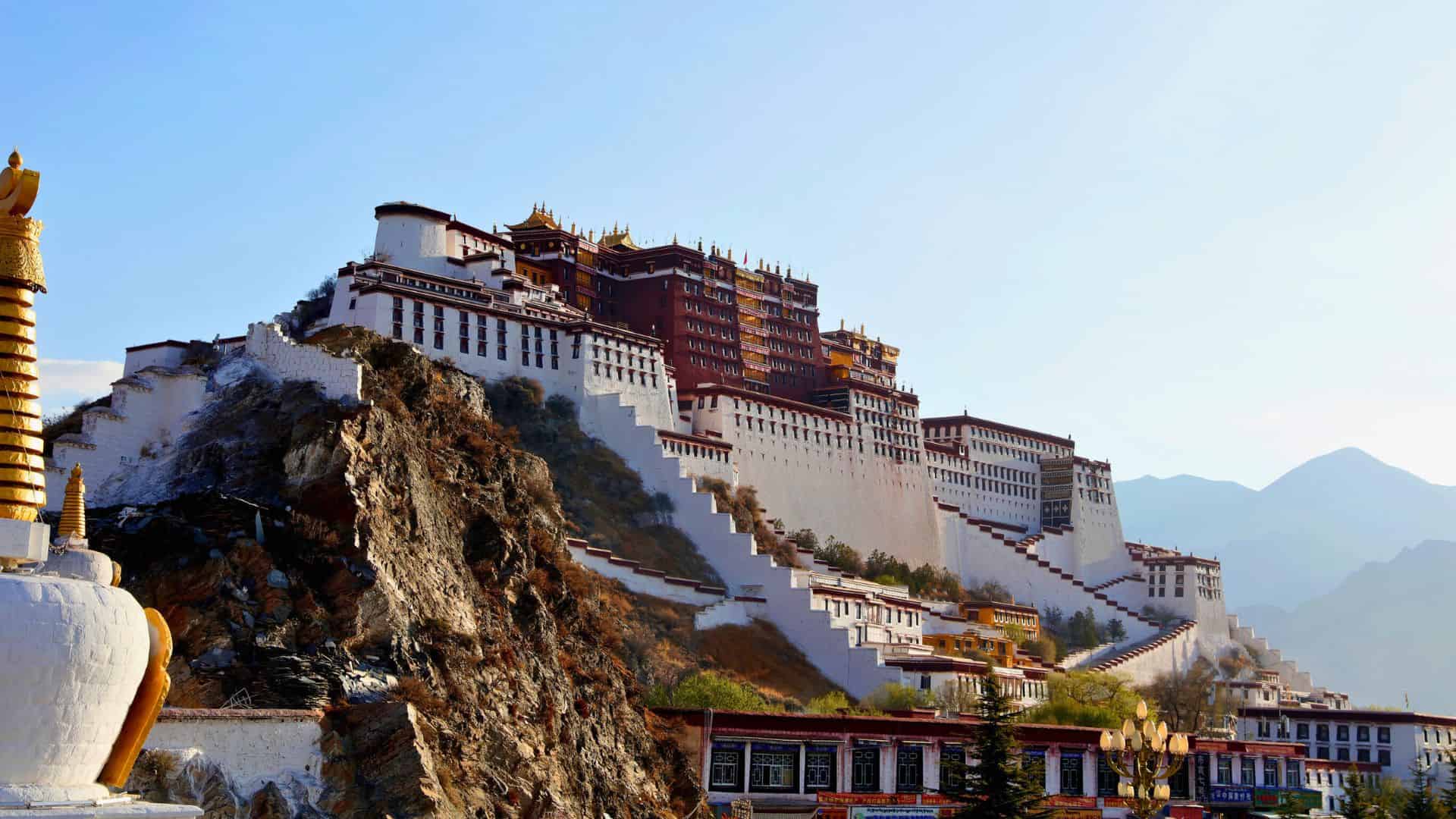 Potala Palace in Lhasa, Tibet, sits atop a rocky hill under a clear blue sky, with mountains in the background.
