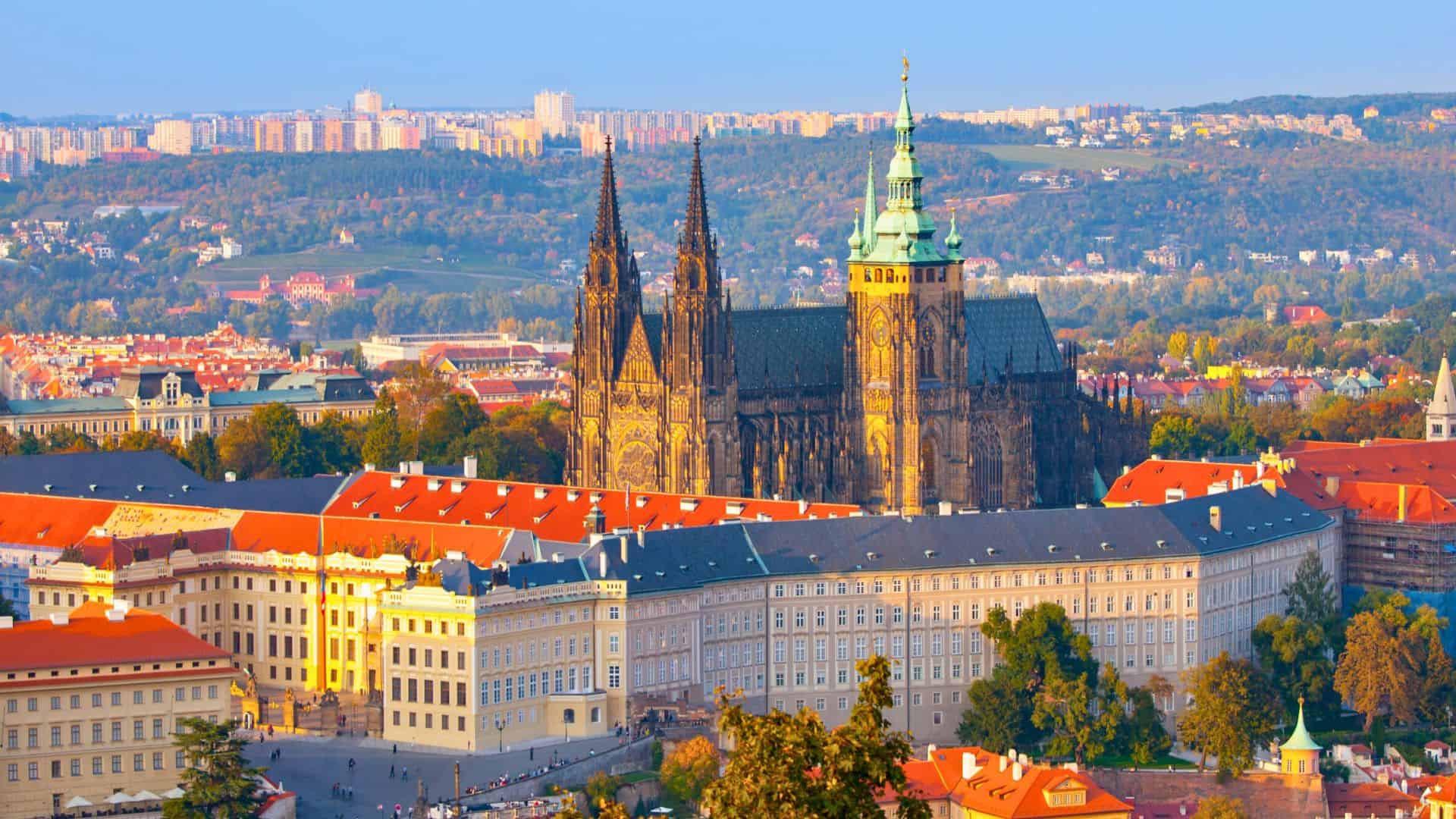 View of Prague Castle and St. Vitus Cathedral with city buildings and trees in the background at sunset.