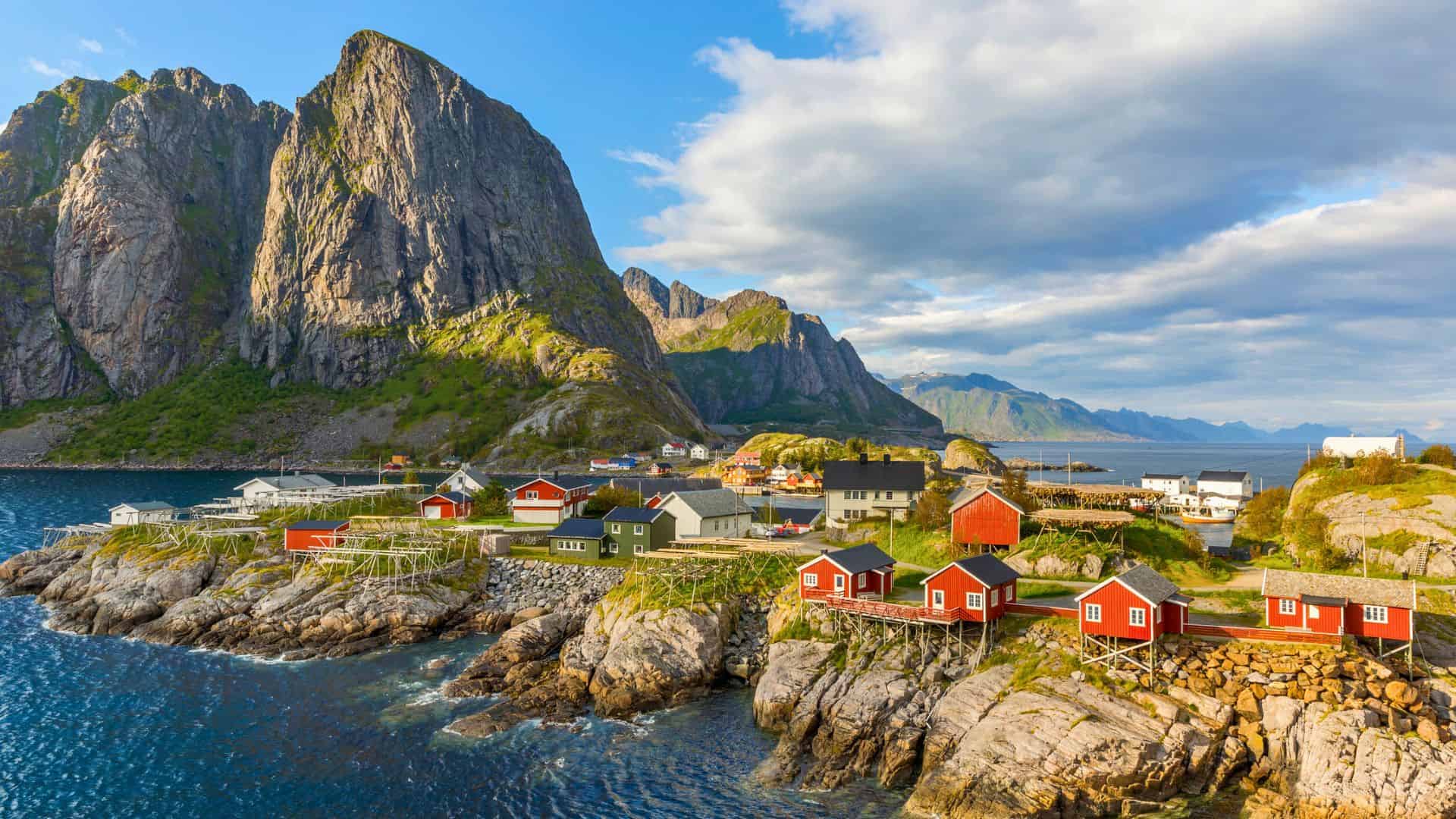 Red cabins on rocky shore with towering mountains and blue water under a partly cloudy sky in a coastal village.
