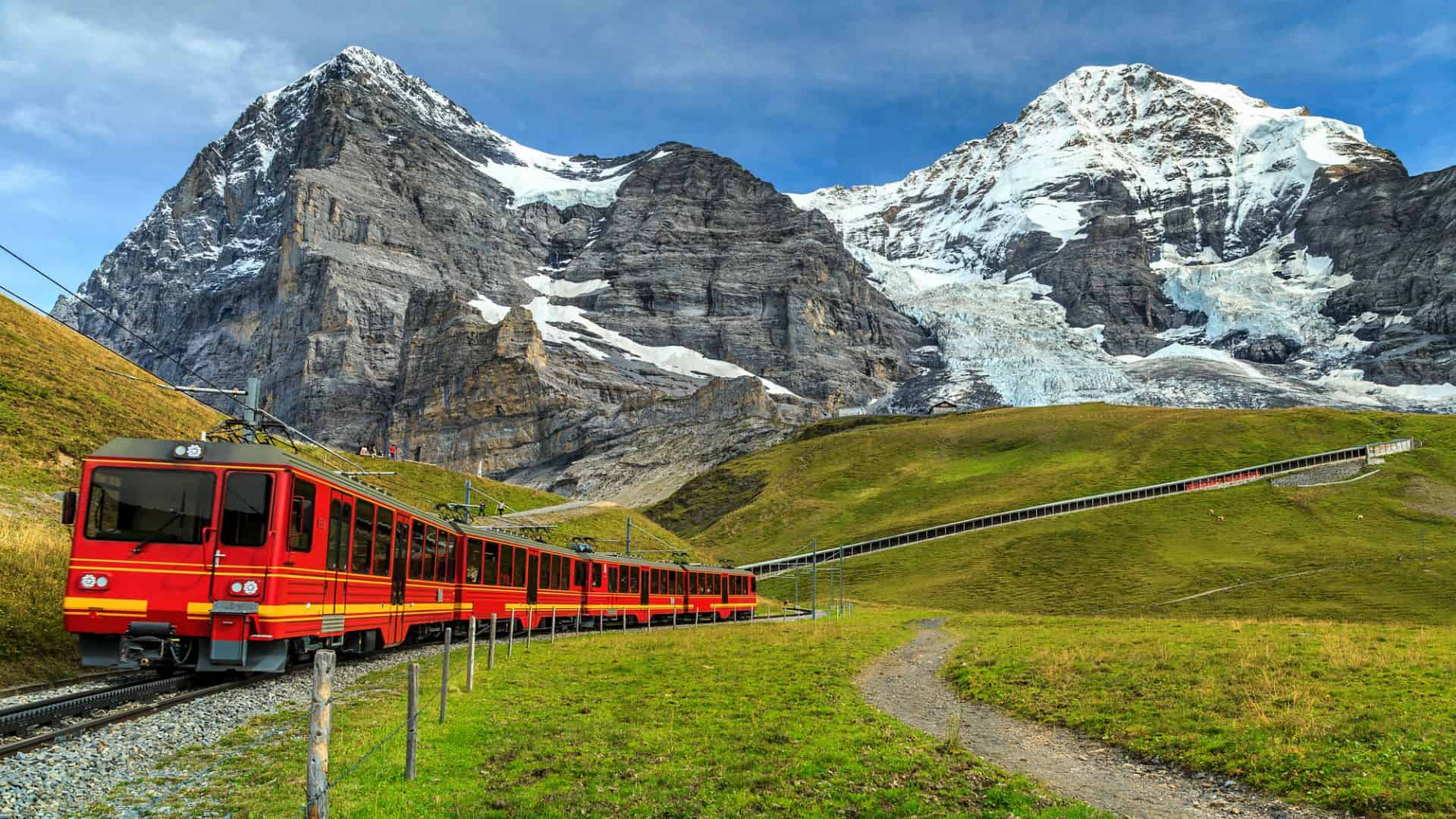 A red and yellow train travels through green hills with snow-capped mountains in the background.