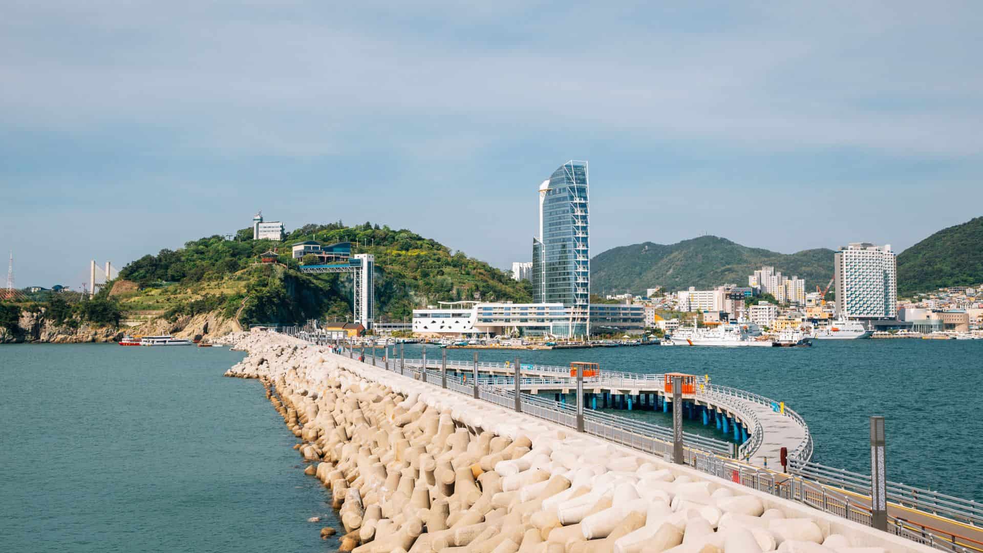 Curved boardwalk over the sea leading to a cityscape with modern buildings and a green hill.