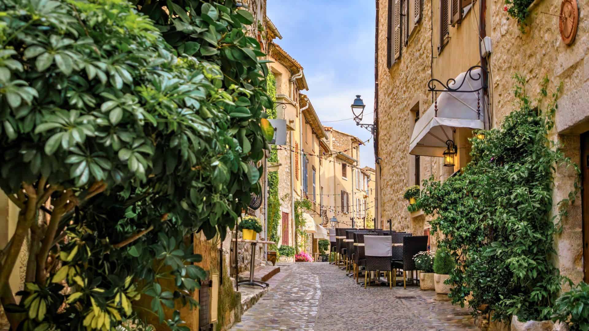 Narrow cobblestone street lined with plants and outdoor caf&eacute; seating between old stone buildings.