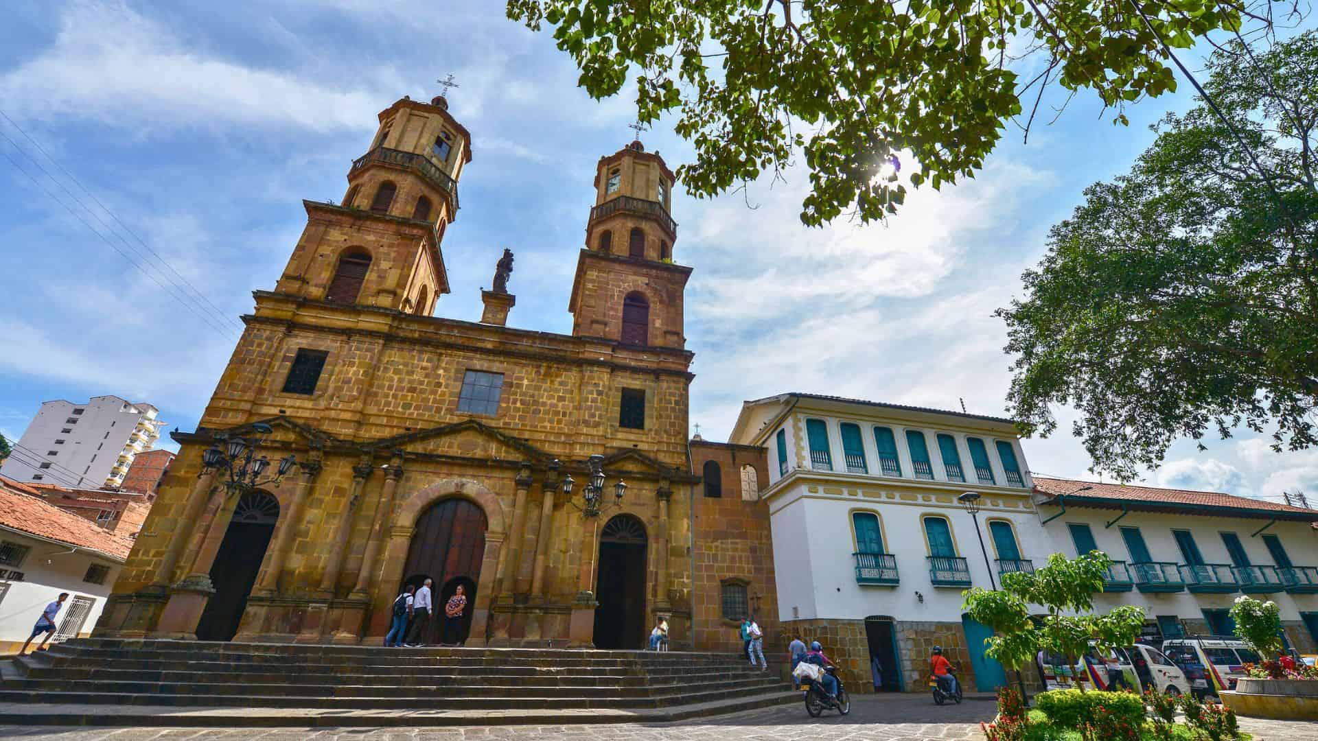 Historic stone church with twin towers beside a colonial-style building, people walking in a sunny plaza.