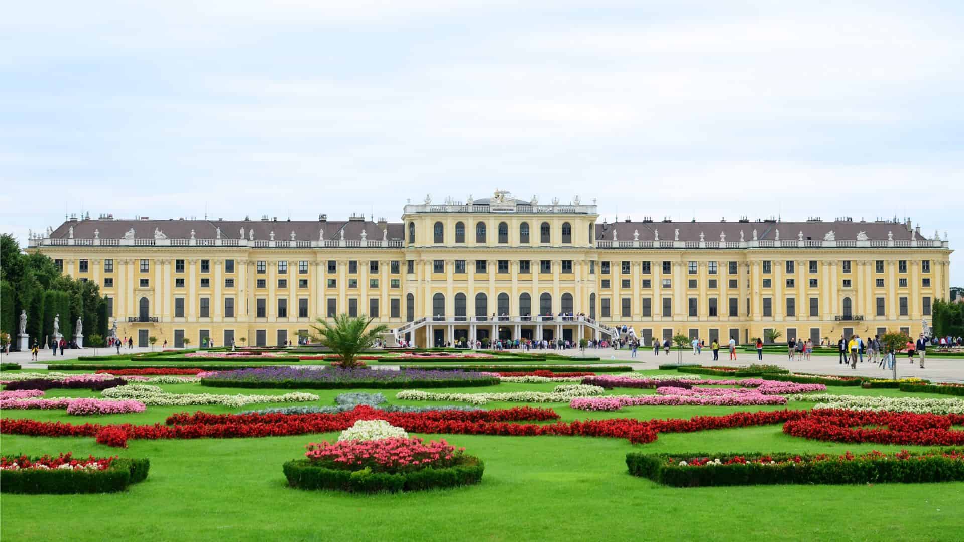 Yellow palace with arched windows and manicured gardens with colorful flowerbeds in the foreground.