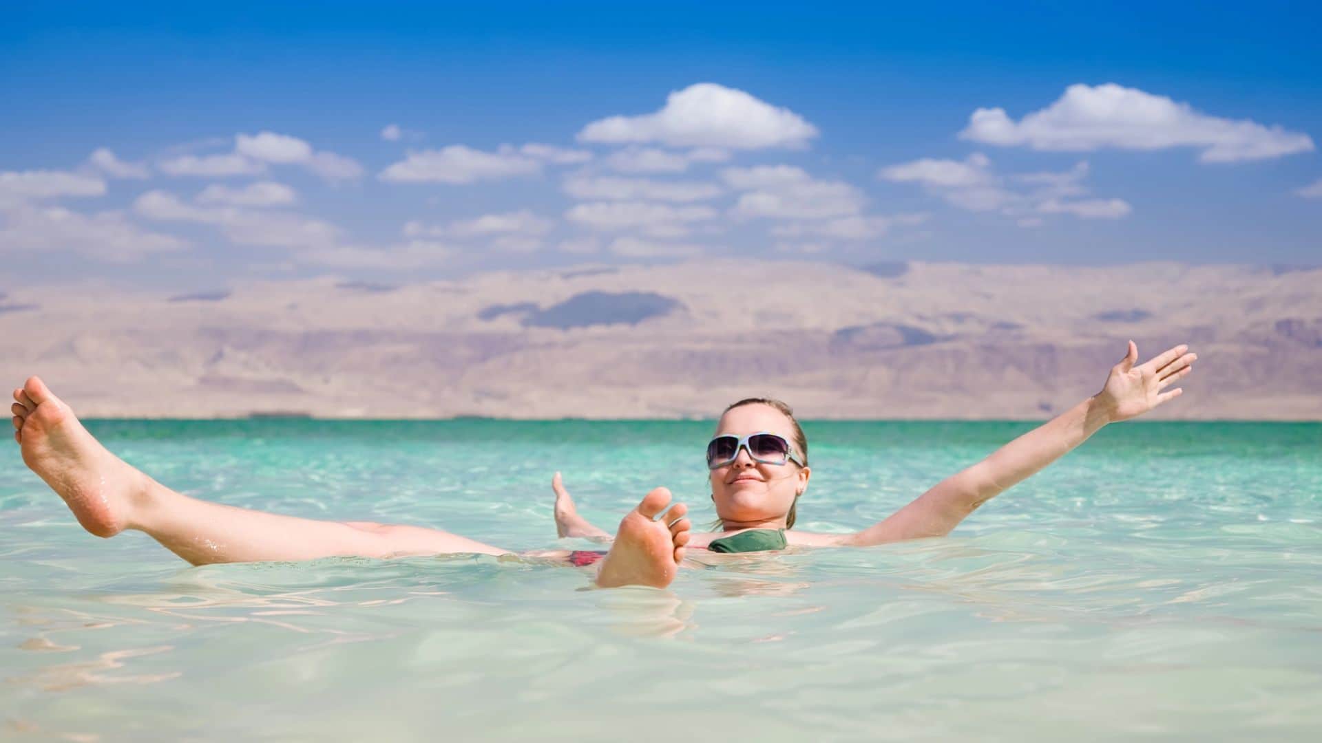 Person floating on their back in clear turquoise water, arms outstretched, with desert mountains in the background.