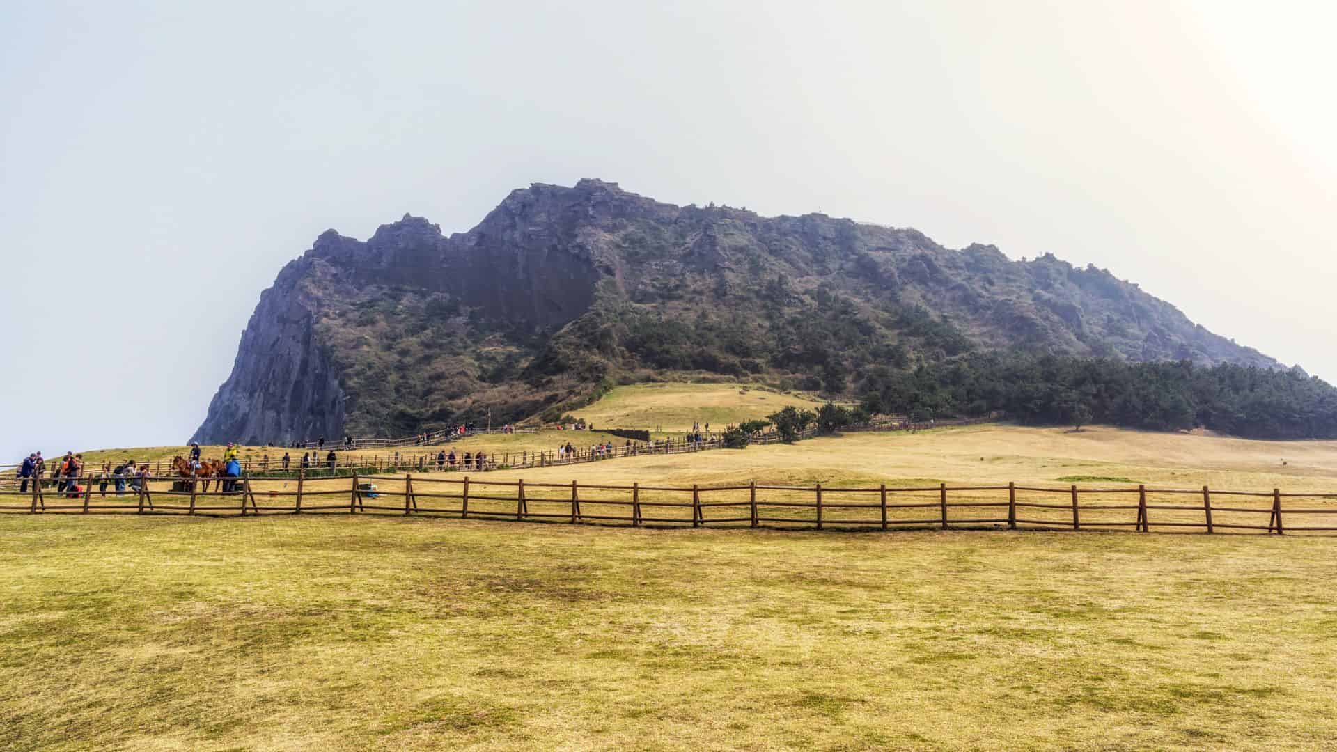 A grassy field with a wooden fence, tourists, and a rocky mountain in the background under a hazy sky.