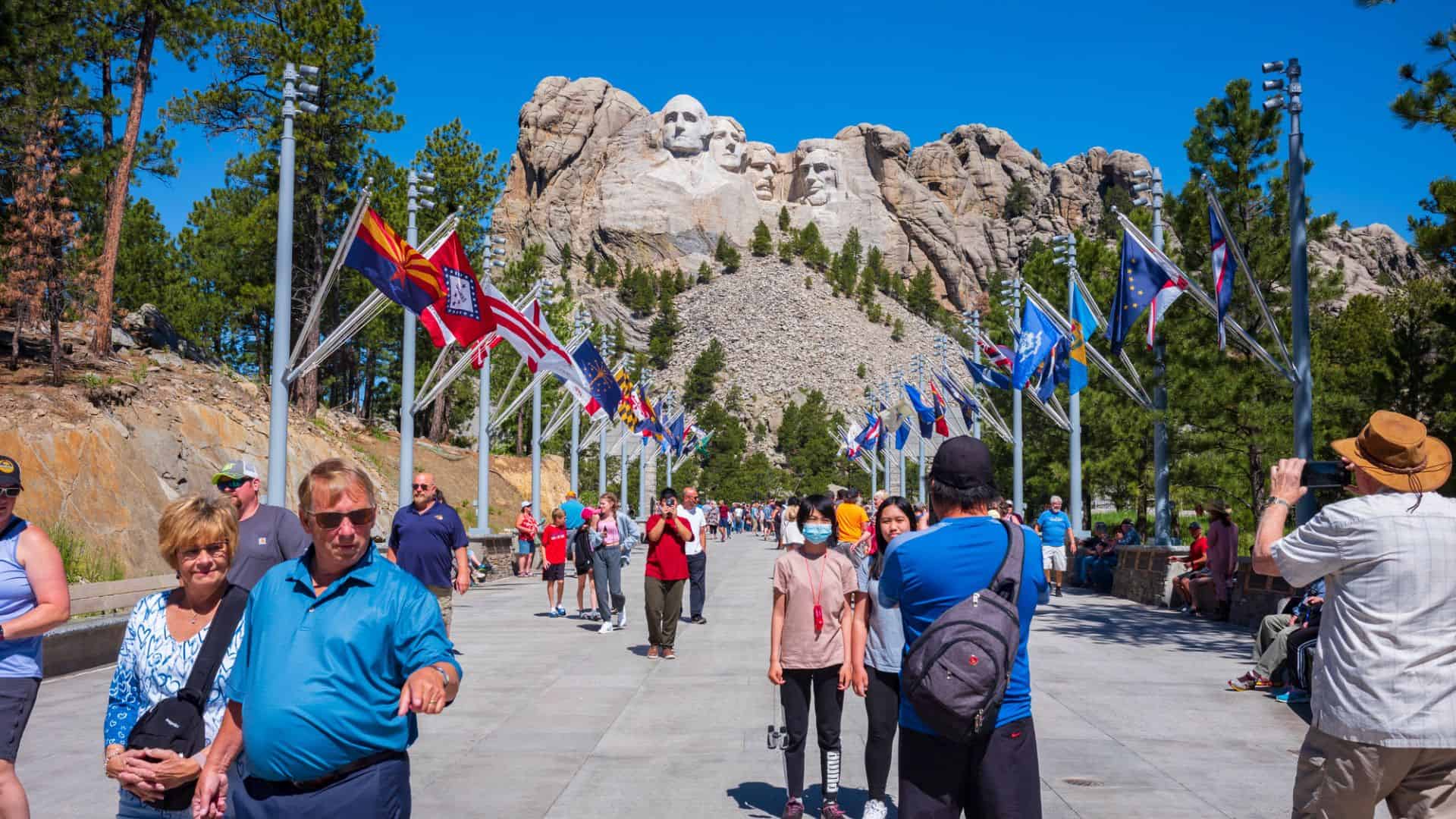 Tourists walk toward Mount Rushmore, flanked by flag poles and trees on a sunny day.