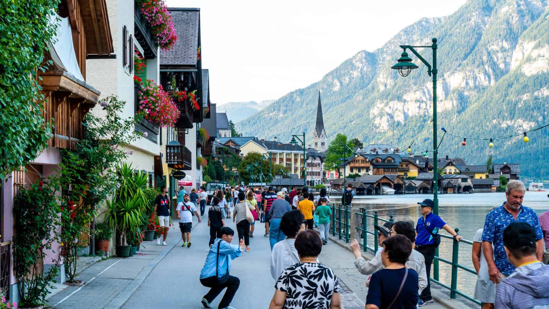 People walking along a lakeside promenade with alpine mountains and colorful buildings in the background.