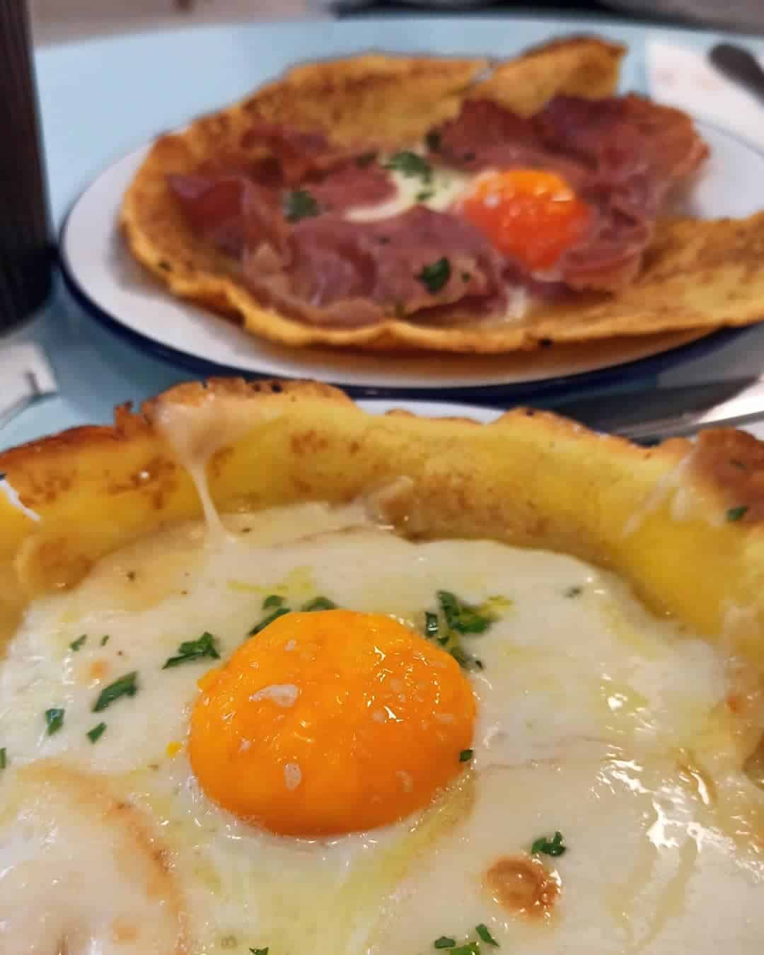 Close-up of two plates with savory pancakes topped with fried eggs and herbs, one with ham slices—perfect for those seeking inspiration from Gluten Free Restaurants in Rome.