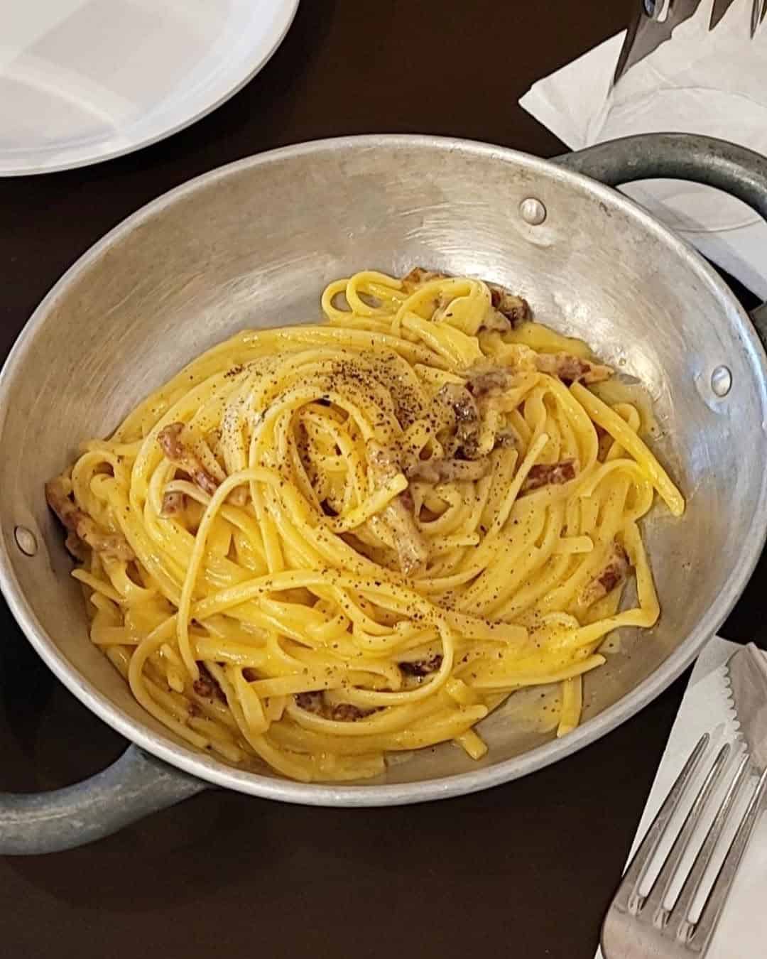A pan of creamy pasta carbonara topped with black pepper, just like you’d find at gluten free restaurants in Rome, placed on a table with utensils nearby.