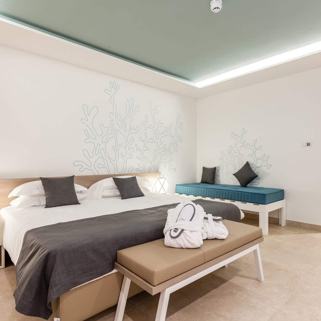 Modern hotel room with a large bed, bench, sofa, wall art, and a folded robe on the bench&mdash;perfect for families seeking comfort at one of the best beach resorts in Italy.