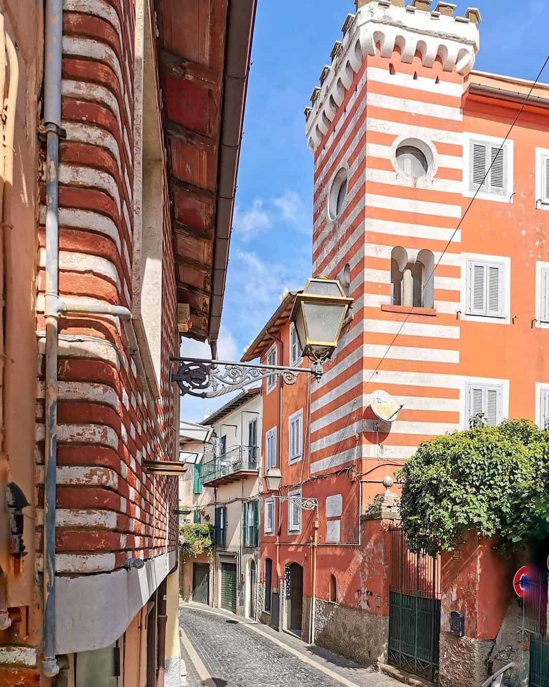 Narrow cobblestone street with striped red and white buildings and a tall tower under a blue sky—perfect for exploring during 4 days in Castel Gandolfo.