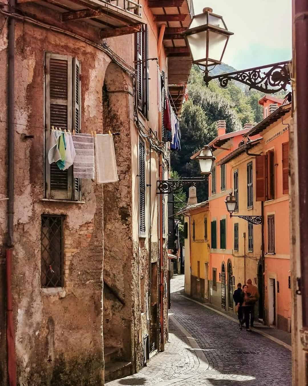 Narrow cobblestone street with old colorful buildings, hanging laundry, and two people walking in sunlight—a charming scene you might encounter during 4 days in Castel Gandolfo.