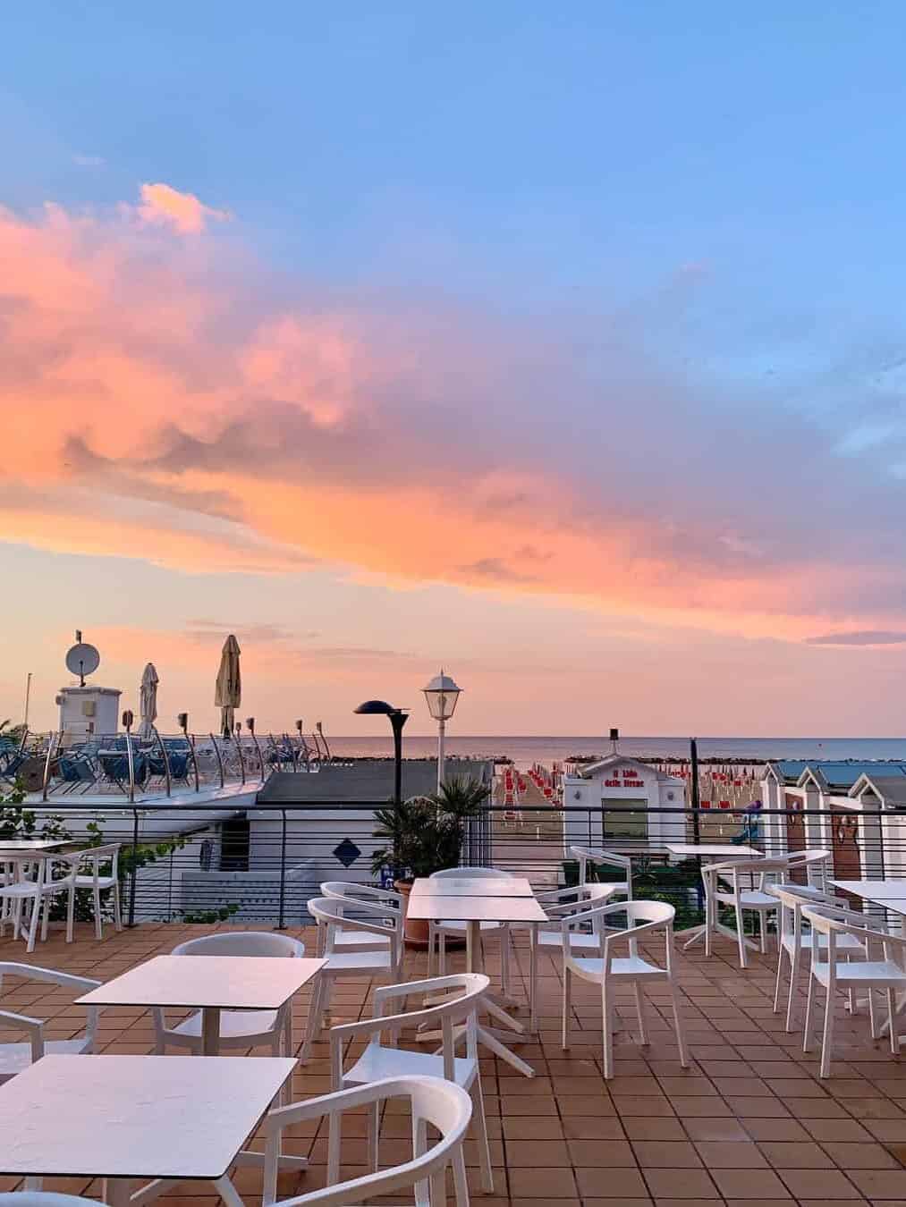 Outdoor rooftop terrace with white chairs and tables, overlooking the sea at sunset with pink and orange clouds&mdash;perfect for relaxing evenings at one of the best beach resorts in Italy for families.