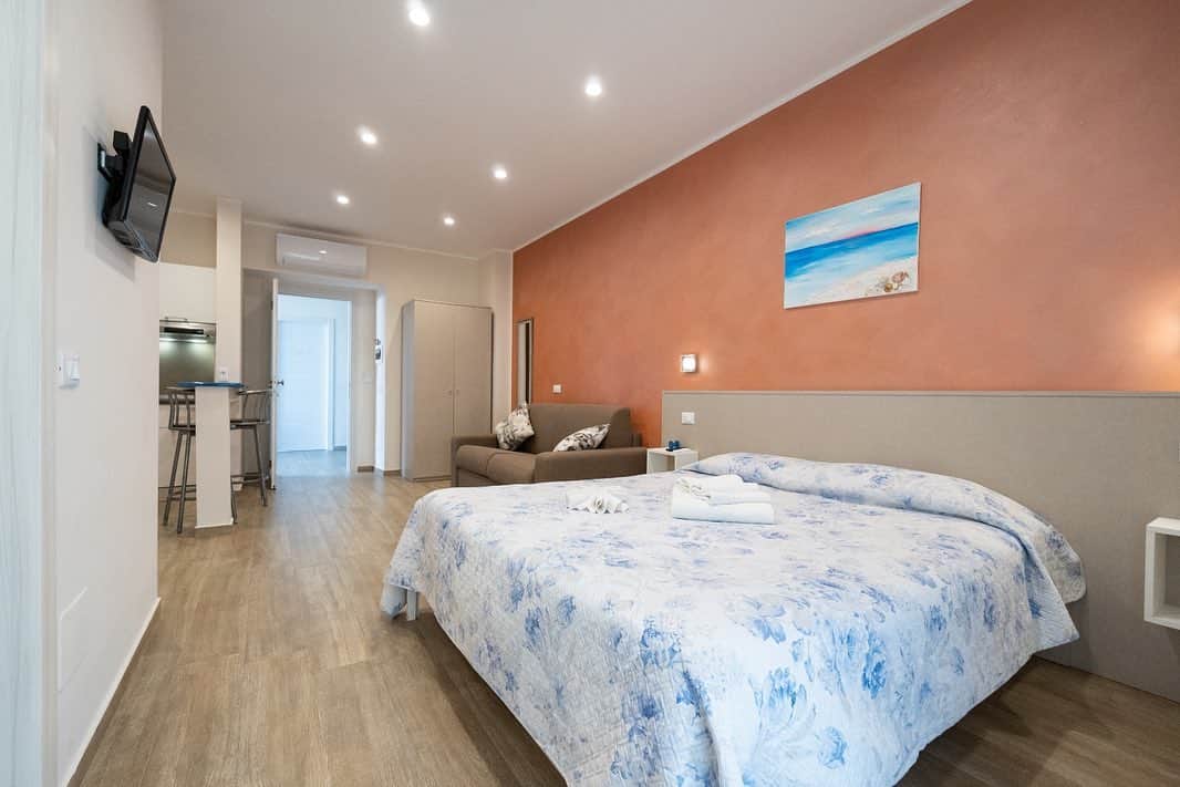 Modern studio with a double bed, sofa, TV, and small kitchen area; peach accent wall with art&mdash;perfect for relaxing after exploring the best beach resorts in Italy for families.