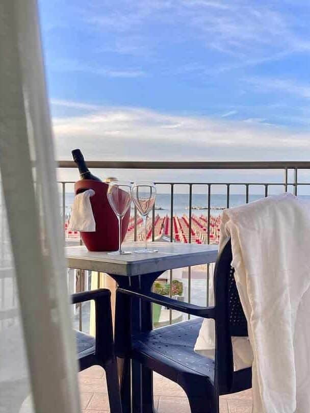 A table on a balcony at one of the best beach resorts in Italy for families, with wine glasses, a bottle chilling in a cooler, and a chair draped with a white towel.