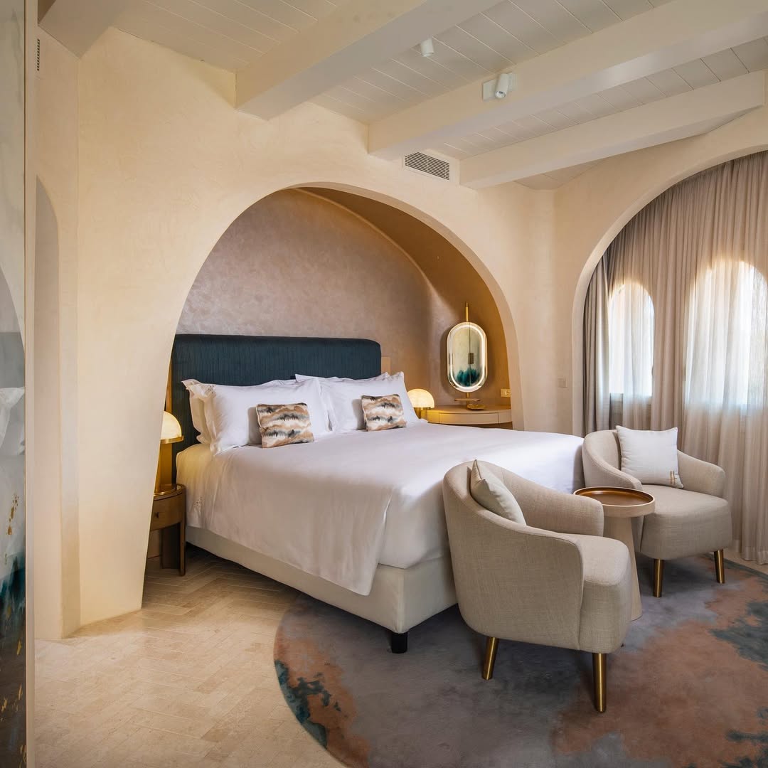Cozy hotel room with a large bed, two armchairs, a round table, arched walls, and soft natural light&mdash;perfect for those seeking the comfort of the best beach resorts in Italy for families.