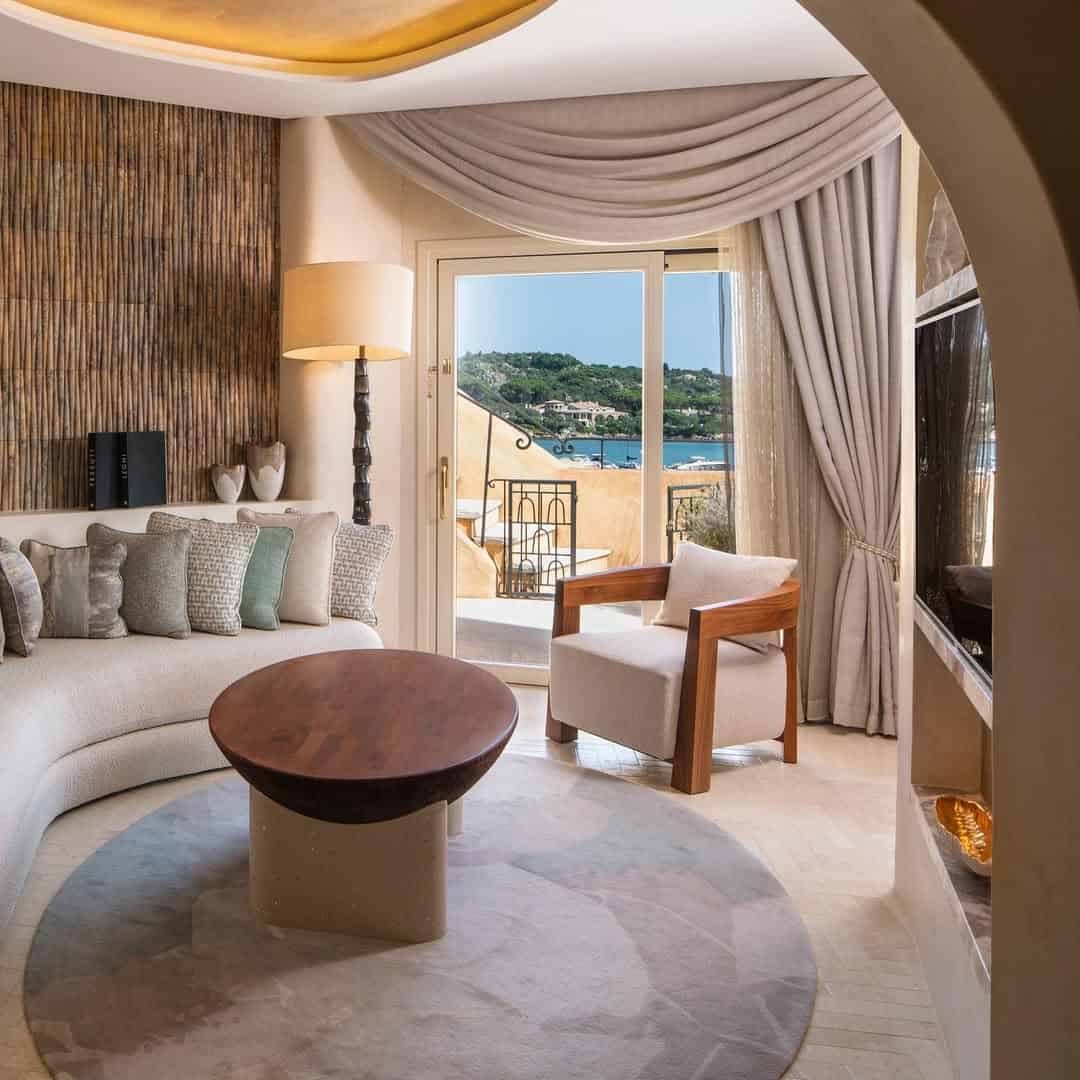 Elegant living room with curved sofa, wooden table, and armchair, overlooking a balcony with a seaside view&mdash;reminiscent of the best beach resorts in Italy for families.