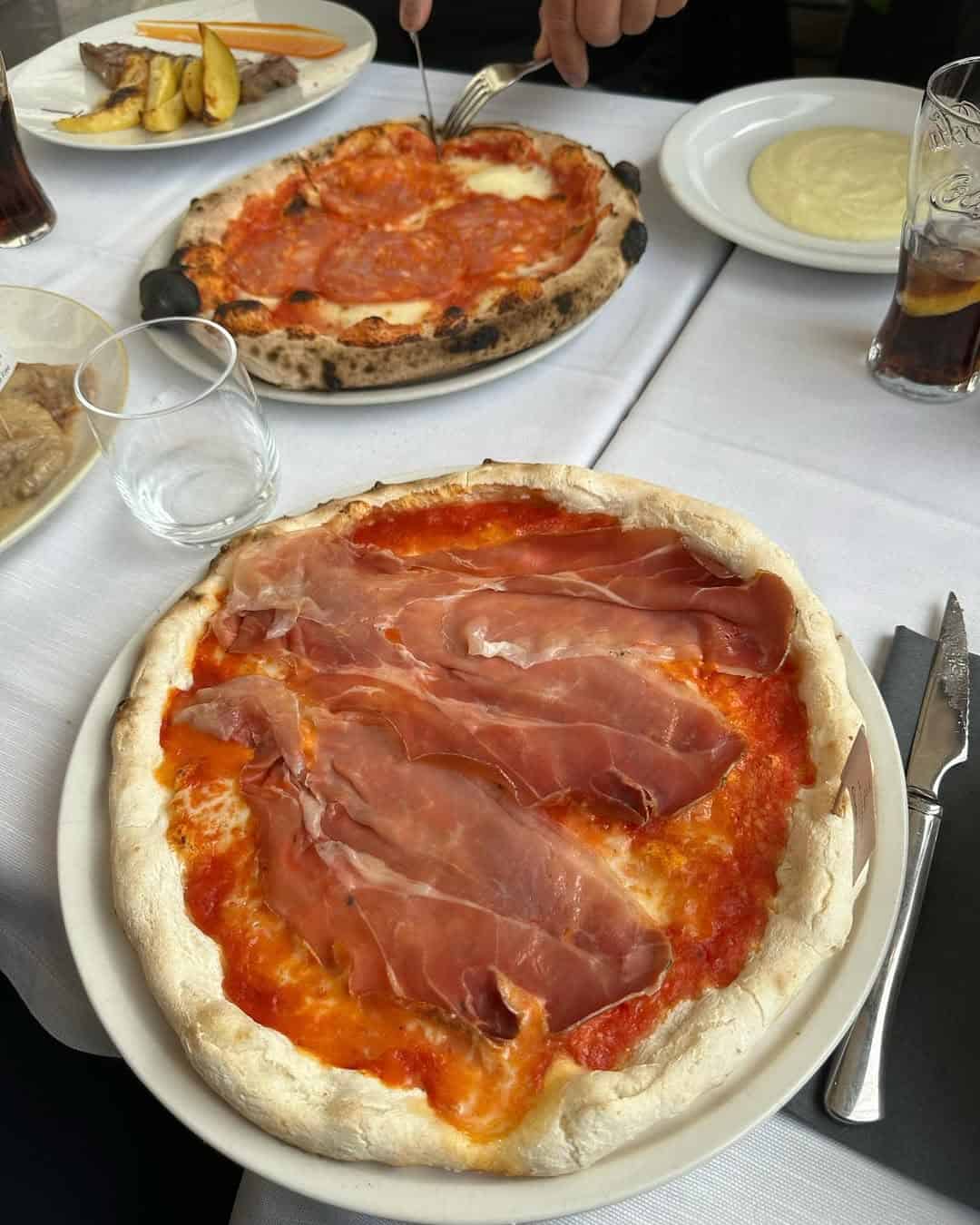 Two pizzas with cured meat toppings on a table set with drinks, plates, and cutlery—a delicious scene you might find at some of the top Gluten Free Restaurants in Rome.