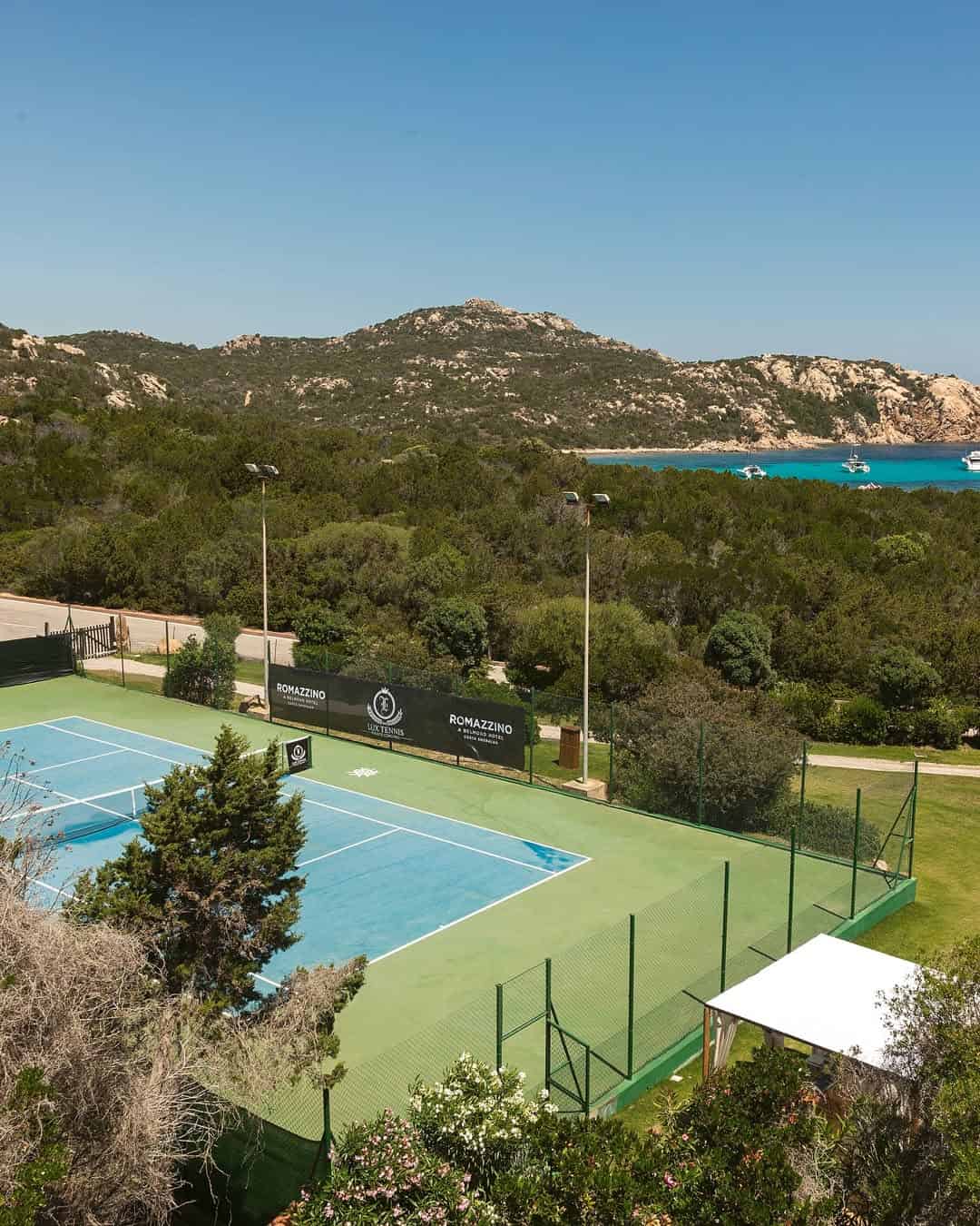 Outdoor tennis court surrounded by trees, with hills and blue sea in the background&mdash;an idyllic setting often found at the best beach resorts in Italy for families, all under a clear sky.