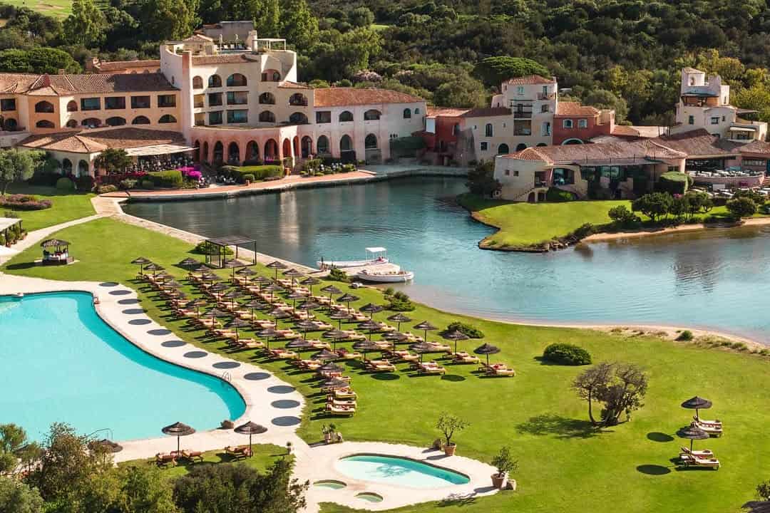 Experience one of the best beach resorts in Italy for families, featuring a large pool, sun loungers, umbrellas, a lagoon, and buildings nestled amid lush greenery.