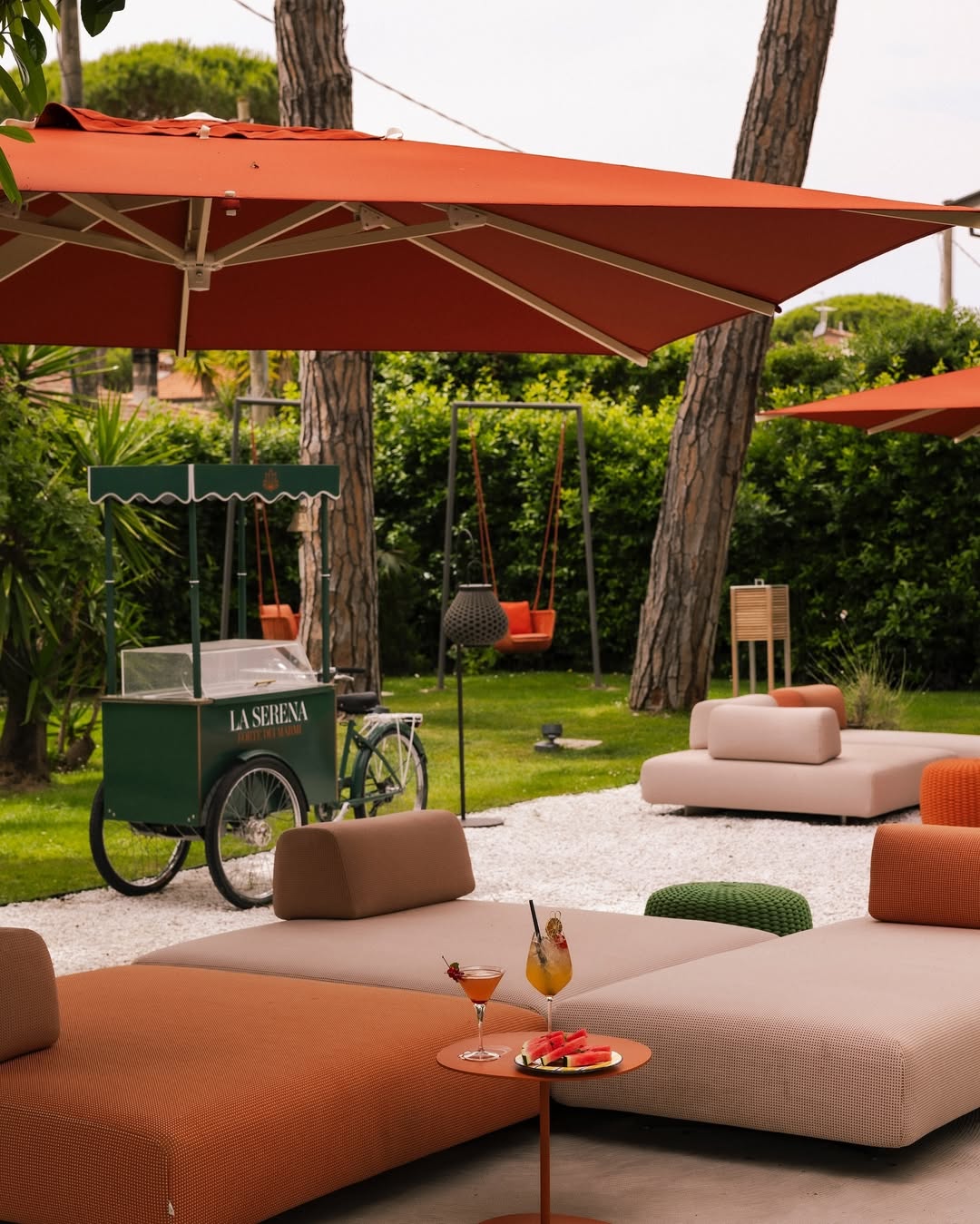 Outdoor lounge with cushioned seating, orange umbrellas, a drink cart, and two cocktails on a table&mdash;just like the relaxing spaces found at the best beach resorts in Italy for families.