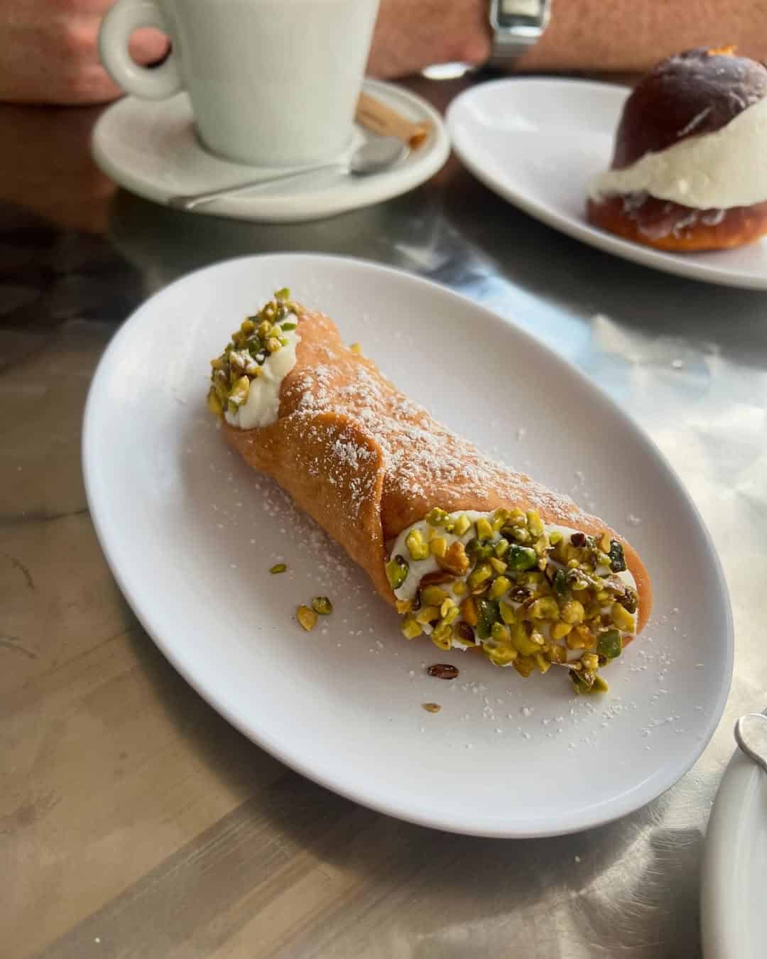 A cannoli with pistachios and powdered sugar on a white plate, echoing the tempting sweets served at top Gluten Free Restaurants in Rome, with coffee and pastry in the background.