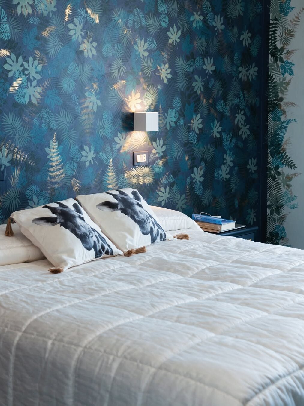 Modern bedroom with a white bed, patterned pillows, and floral blue wallpaper&mdash;evoking the relaxing vibes found at the best beach resorts in Italy for families.