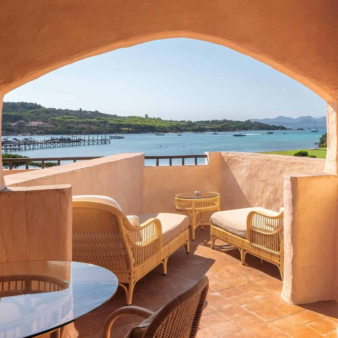 Sunny terrace with wicker chairs and a table overlooking a bay, pier, and green hills&mdash;an inviting spot at one of the best beach resorts in Italy for families.