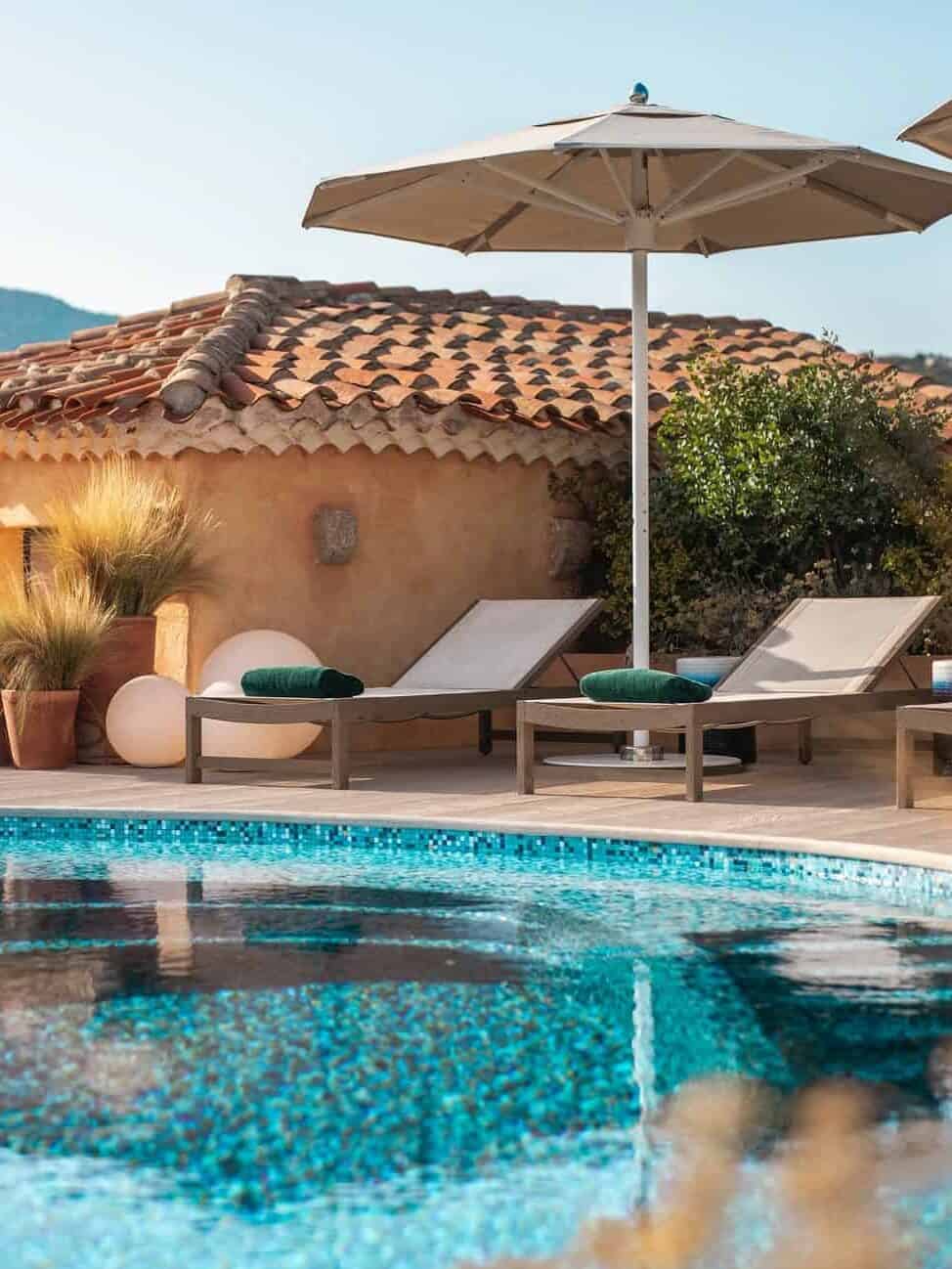 Two lounge chairs and an umbrella by a pool evoke the relaxing atmosphere found at the best beach resorts in Italy for families, with a tiled-roof building and lush plants in the background.
