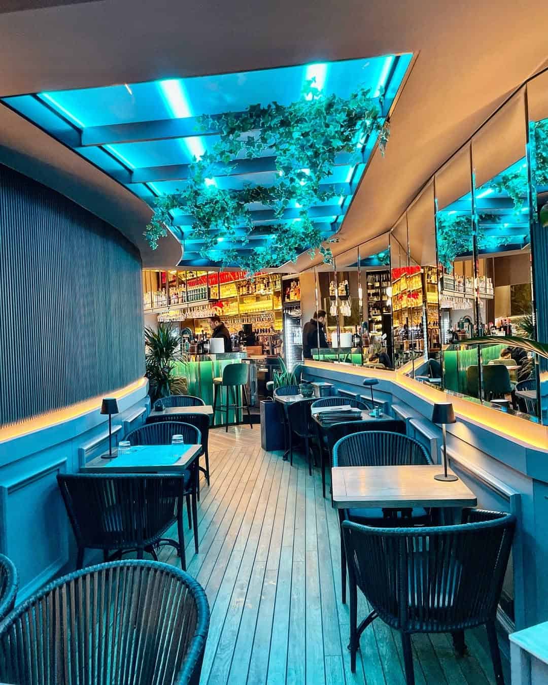 Modern bar with blue ceiling lights, hanging plants, wooden floors, and tables with black chairs—perfect for those seeking stylish Gluten Free Restaurants in Rome.