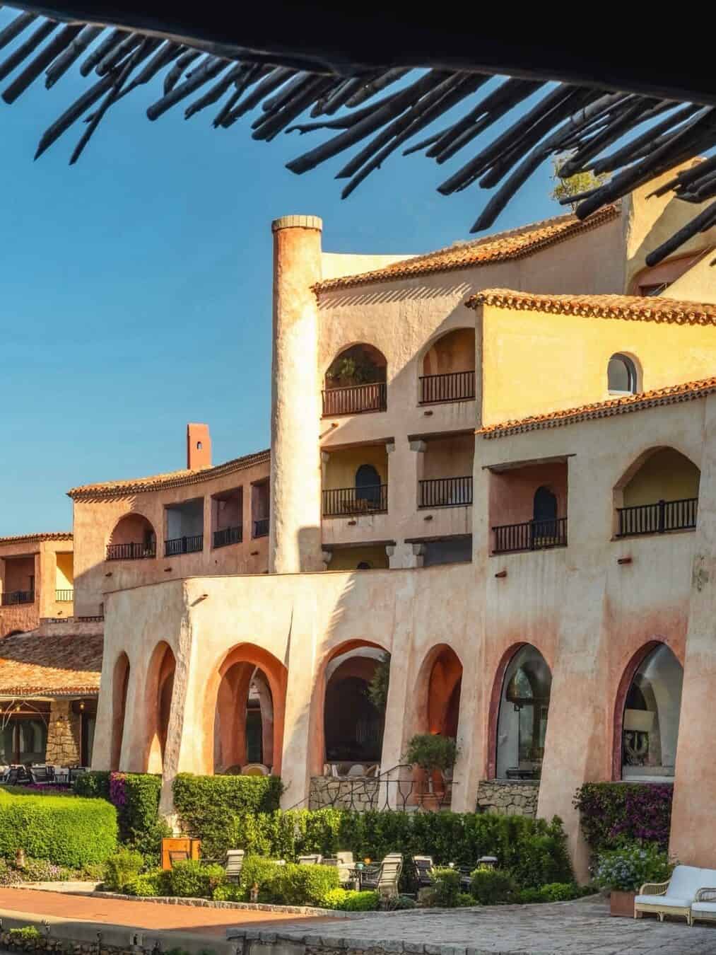 A sunlit Mediterranean-style hotel building with arches, balconies, and lush landscaping in front&mdash;perfect for families seeking one of the best beach resorts in Italy.