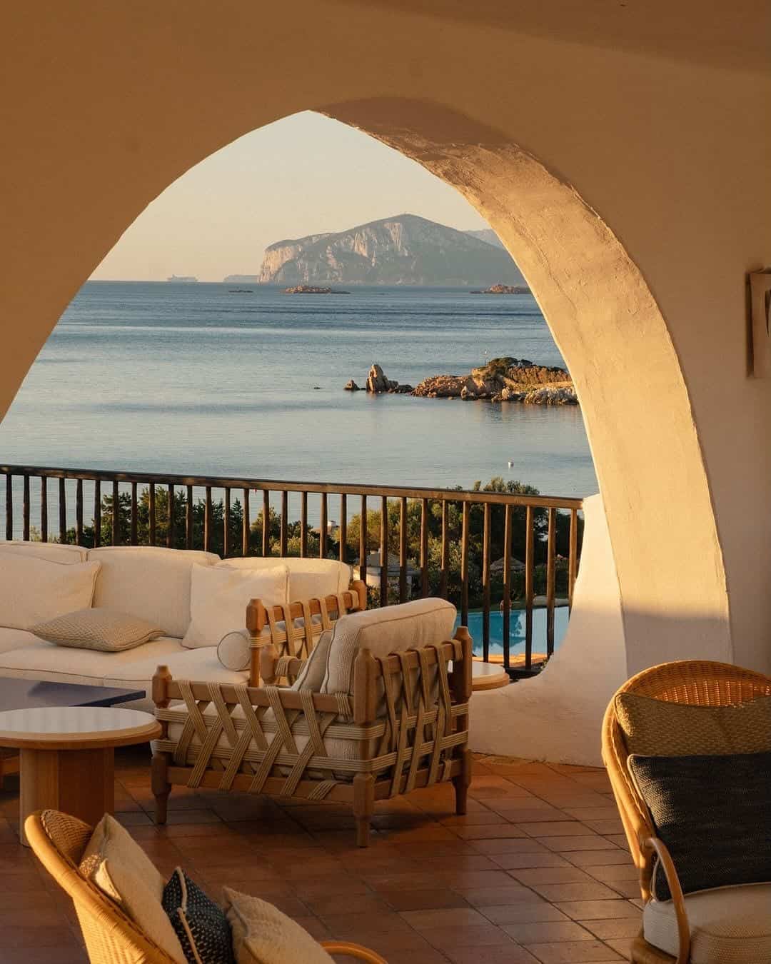A cozy patio with wicker furniture overlooks a calm sea and distant cliffs through a large arched opening, capturing the charm found at the best beach resorts in Italy for families.
