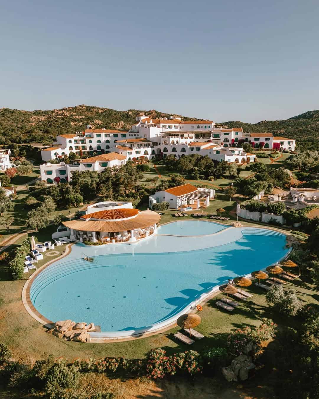 A large outdoor pool with sunbeds in front of a hillside resort, surrounded by greenery and mountains&mdash;perfect for those seeking the best beach resorts in Italy for families.