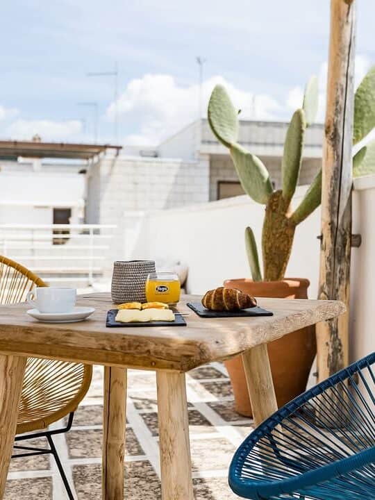 Breakfast with coffee and croissants on a patio table with two chairs and a potted cactus, under a blue sky&mdash;reminiscent of mornings at the best beach resorts in Italy for families.